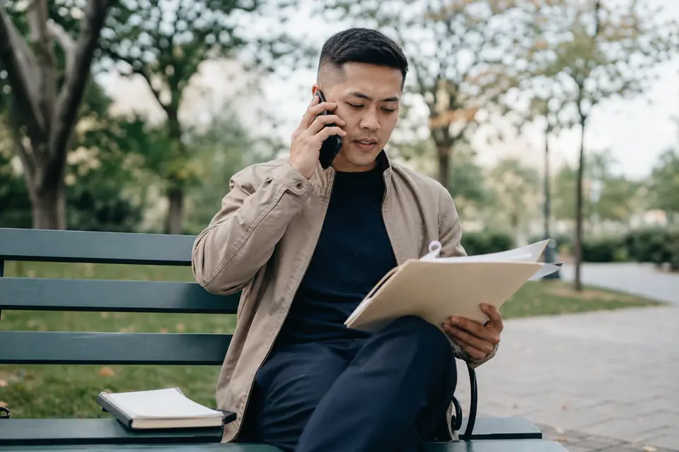 Young man on a park bench on a phone call with tax papers on his lap