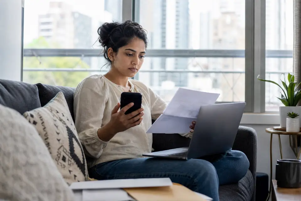 Young woman on a couch reviewing a letter while using a laptop and phone
