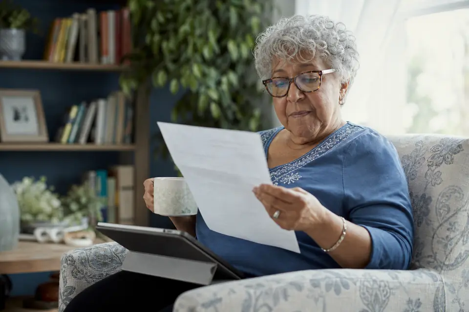 Older woman reading a one-page letter in an armchair with a tablet